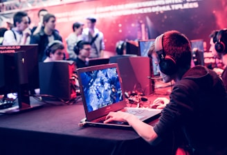 Several people are seated and focused on computer screens, wearing headsets, in an environment that appears to be a gaming tournament or esports event. The room is dimly lit with neon lighting, highlighting screens and creating an intense atmosphere.