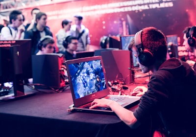 Several people are seated and focused on computer screens, wearing headsets, in an environment that appears to be a gaming tournament or esports event. The room is dimly lit with neon lighting, highlighting screens and creating an intense atmosphere.
