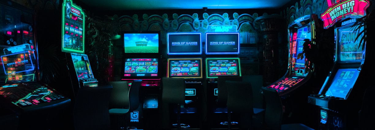 Dimly lit gaming arcade featuring a variety of brightly illuminated slot machines and video gaming screens. Neon signs and colorful displays create a vibrant atmosphere. The arrangement of gaming machines suggests an inviting space for entertainment, with chairs positioned for players.