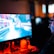 A person wearing headphones is sitting in front of a computer monitor. The screen displays a vibrant, illuminated video game scene, while the person appears focused on gaming. The ambient lighting is dark with red and blue tones, and a backlight from the monitor highlights the headphones.