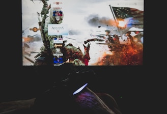 A dimly lit room with a television screen displaying a dynamic, action-packed video game interface featuring a historical battle scene. The foreground shows the silhouette of a gaming console controller held by a person.