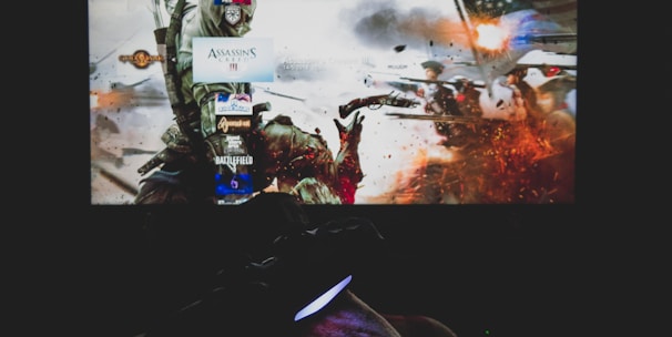 A dimly lit room with a television screen displaying a dynamic, action-packed video game interface featuring a historical battle scene. The foreground shows the silhouette of a gaming console controller held by a person.