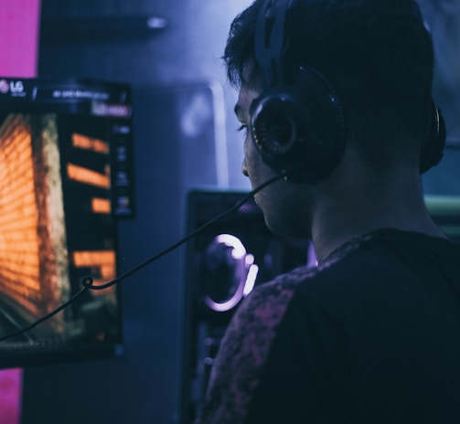 A person wearing headphones is engaged in gaming on a computer, with a large screen displaying a video game scene, showing a wooden building and a path.