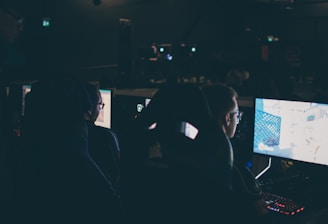 Several individuals are sitting in front of computer screens in a dimly lit room, likely engaging in gaming or working on tasks. The glow of the monitors illuminates their faces, creating a focused and immersive atmosphere. The surroundings suggest a setting like a gaming event or a LAN party.
