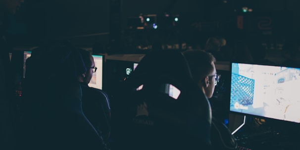 Several individuals are sitting in front of computer screens in a dimly lit room, likely engaging in gaming or working on tasks. The glow of the monitors illuminates their faces, creating a focused and immersive atmosphere. The surroundings suggest a setting like a gaming event or a LAN party.