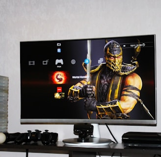 A flat-screen TV displays a video game interface featuring a character from Mortal Kombat in the center. Beside the TV is a stack of video game cases for the PlayStation 3, including titles like FIFA 14 and Grand Theft Auto. Below the TV, there is a gaming console, controllers, and other gaming equipment.