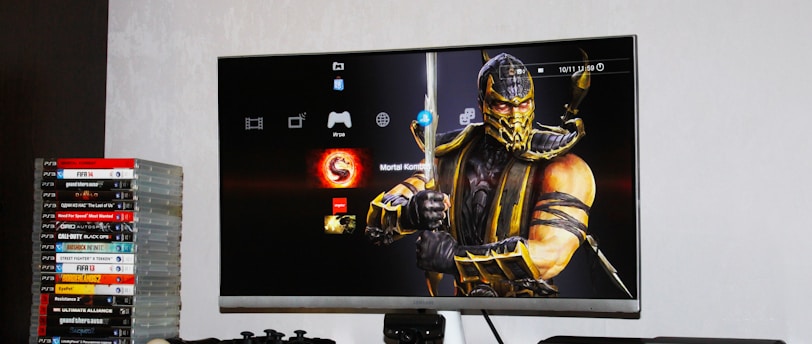 A flat-screen TV displays a video game interface featuring a character from Mortal Kombat in the center. Beside the TV is a stack of video game cases for the PlayStation 3, including titles like FIFA 14 and Grand Theft Auto. Below the TV, there is a gaming console, controllers, and other gaming equipment.