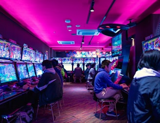 A vibrant arcade with rows of gaming machines adorned with colorful screens. Several people are immersed in playing games, seated on stools in a dimly lit room accented by an intense pink ceiling light. The atmosphere is lively and engaging.