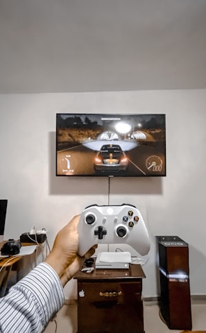 A person is holding a gaming controller in front of a flat-screen television displaying a racing video game. The room has a computer setup on a wooden desk with cables, a desktop computer monitor, and other accessories. The focus is on the interaction between the person and the gaming environment.