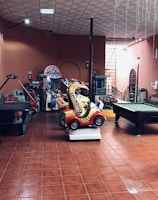 An indoor arcade area featuring various gaming machines, including an air hockey table and a ride-on toy car. The room has a tiled floor and several colorful gaming devices scattered around. Chairs and arcade signage are visible, creating a playful and entertainment-focused atmosphere.