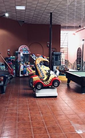 An indoor arcade area featuring various gaming machines, including an air hockey table and a ride-on toy car. The room has a tiled floor and several colorful gaming devices scattered around. Chairs and arcade signage are visible, creating a playful and entertainment-focused atmosphere.