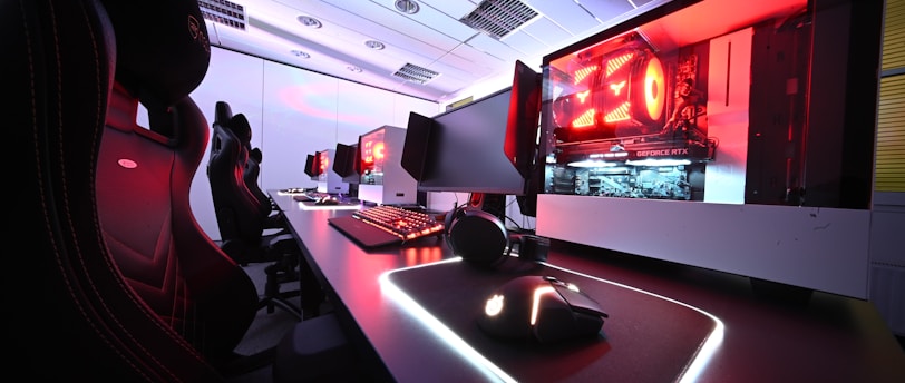 A row of high-end gaming computers on sleek desks, featuring RGB lighting and gaming accessories. The computers are housed in transparent cases with visible components, emitting a red glow. Comfortable gaming chairs with ergonomic design are positioned next to the desks, and the room's ceiling is modern with recessed lighting.
