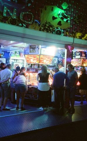 A group of people standing in front of an illuminated amusement arcade, engaging with various gaming machines. The environment is bustling with activity, and colorful lights create a vibrant atmosphere. The ceiling is decorated with large, playful images of dice and other game-related graphics, enhancing the lively mood.