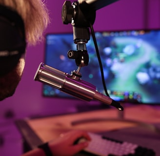 A person wearing headphones is seated in front of a computer screen, engaged in a video game. A microphone is positioned prominently in front of them, suggesting a streaming or recording setup. The background is illuminated with soft purple lighting, creating a cozy and immersive atmosphere.