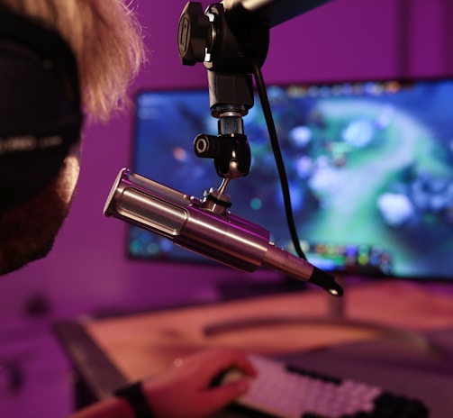 A person wearing headphones is seated in front of a computer screen, engaged in a video game. A microphone is positioned prominently in front of them, suggesting a streaming or recording setup. The background is illuminated with soft purple lighting, creating a cozy and immersive atmosphere.