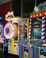 A row of brightly colored arcade game machines is arranged in a line against the wall. Each machine features vibrant lights and designs, with themes ranging from claw games to game show-inspired setups. The area has a playful and inviting atmosphere, suitable for entertainment or amusement centers.