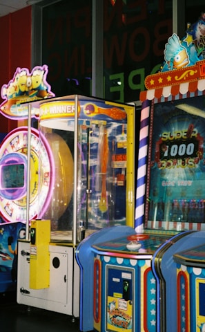 A row of brightly colored arcade game machines is arranged in a line against the wall. Each machine features vibrant lights and designs, with themes ranging from claw games to game show-inspired setups. The area has a playful and inviting atmosphere, suitable for entertainment or amusement centers.
