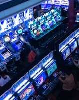 A bustling arcade filled with rows of gaming machines, each with vibrant displays and controls. Several people are seated at these machines, deeply engaged in playing games. Bright fluorescent lights cast a blue and neon glow across the room, enhancing the lively and energetic atmosphere.