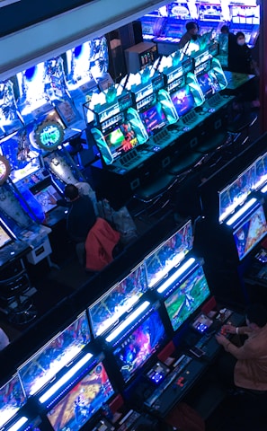A bustling arcade filled with rows of gaming machines, each with vibrant displays and controls. Several people are seated at these machines, deeply engaged in playing games. Bright fluorescent lights cast a blue and neon glow across the room, enhancing the lively and energetic atmosphere.
