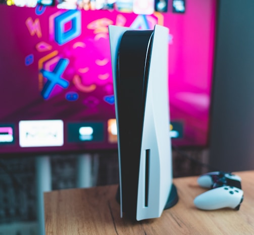 A gaming console is placed on a wooden table with a game controller nearby. In the background, a television screen displays colorful graphics, including geometric shapes and vibrant colors.