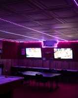 A dimly lit room with vibrant purple LED strip lights on the ceiling, featuring multiple gaming stations. There are two large screens showing video games, along with various arcade machines, including a Nintendo arcade cabinet. An air hockey table is positioned in the foreground.