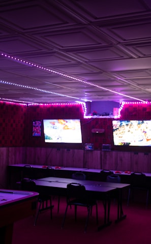 A dimly lit room with vibrant purple LED strip lights on the ceiling, featuring multiple gaming stations. There are two large screens showing video games, along with various arcade machines, including a Nintendo arcade cabinet. An air hockey table is positioned in the foreground.
