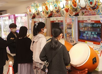 Several people are engaged with arcade games, focusing on rhythm games with large drum controllers. The players appear focused and are wearing masks, indicating a casual, indoor setting in an arcade filled with vibrant colors.