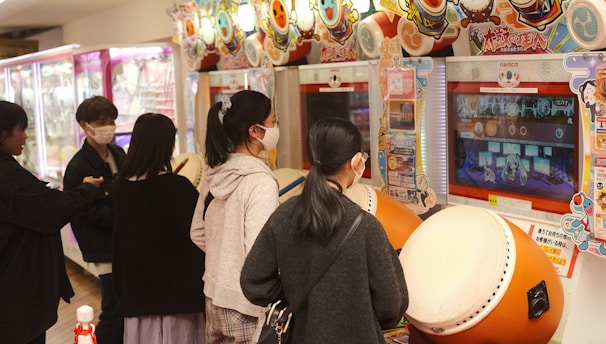 Several people are engaged with arcade games, focusing on rhythm games with large drum controllers. The players appear focused and are wearing masks, indicating a casual, indoor setting in an arcade filled with vibrant colors.