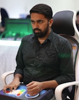 A person is seated in a chair, focused intently on something, with hands positioned on a gaming console or arcade controller. The setup appears to be indoors, with visible screens and lighting in the background, suggesting an electronic gaming environment.