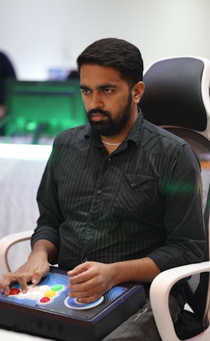 A person is seated in a chair, focused intently on something, with hands positioned on a gaming console or arcade controller. The setup appears to be indoors, with visible screens and lighting in the background, suggesting an electronic gaming environment.